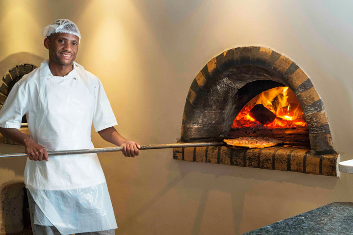 Smiling chef holding a pizza peel by a stone wood-fired oven with visible flames