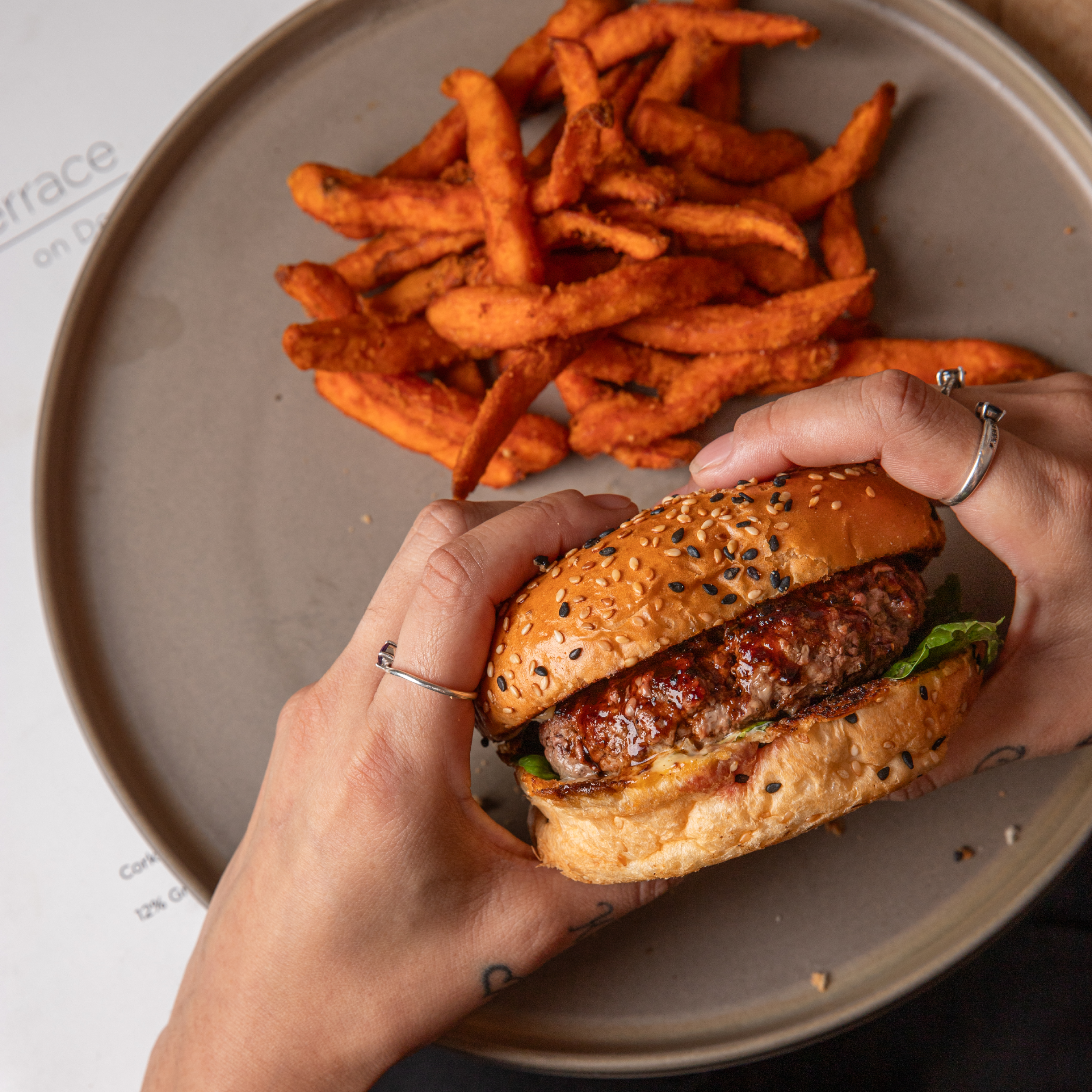Hands holding sesame-seed burger above plate of sweet potato fries