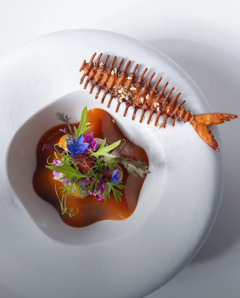Crispy fried fish skeleton next to amber broth topped with edible flowers