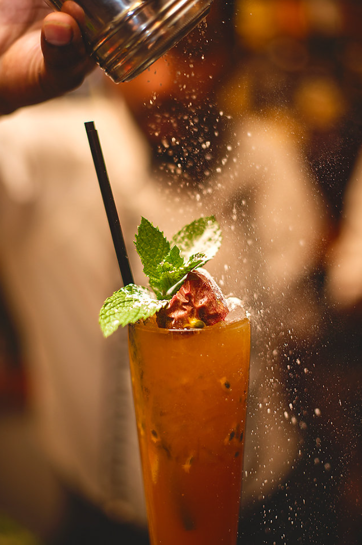 Hand sprinkling powder over tall orange cocktail garnished with mint