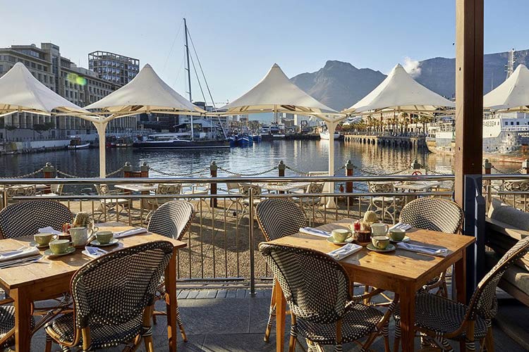 Outdoor waterfront café tables with cups overlooking a marina with white canopy tents and boats