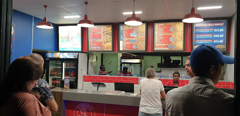 Customers queue at Roman's Pizza Durbanville counter with staff and lit menu boards