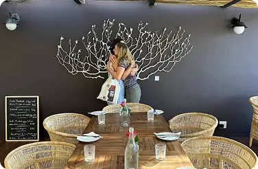 Two women hugging beneath white coral mural in dining area