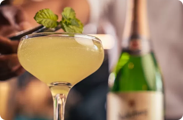 Pale yellow cocktail in a coupe with mint garnish beside a blurred sparkling-wine bottle